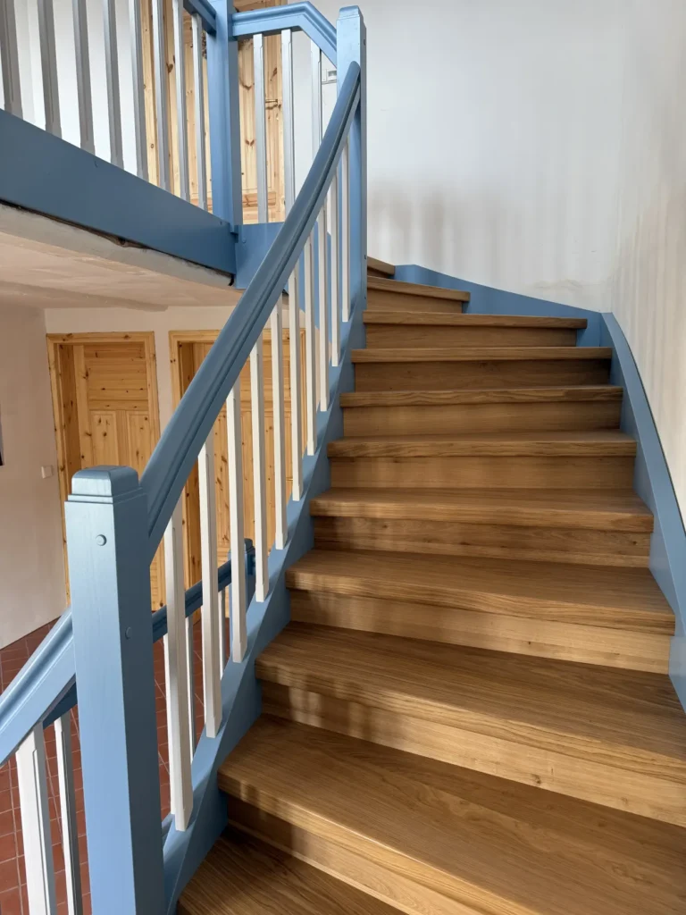 Eine Holztreppe mit hellblauen Geländern und weißen Balustern schlängelt sich neben einer weißen Wand nach oben. Unten links sind Türen mit Holzpaneelen und ein rot gekachelter Boden zu sehen.