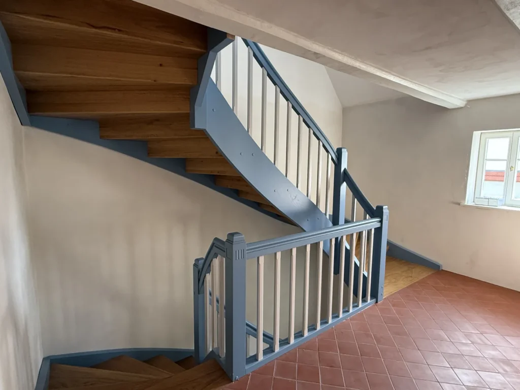Eine Holztreppe mit hellblauen Geländern und weißen Balustern schlängelt sich in einer Ecke eines Raums mit Fliesenboden und einem Fenster, durch das Tageslicht hereinfällt, nach oben.