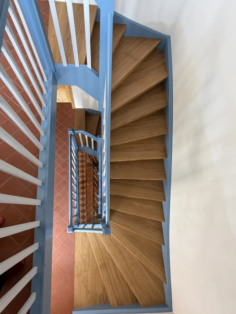 Eine Ansicht von oben auf eine Holztreppe mit blauen Geländern, die an einem Treppenabsatz eine scharfe Kurve macht. Die Treppe führt hinunter zu einem Boden mit terrakottafarbenen Fliesen, die in einem Rautenmuster verlegt sind.