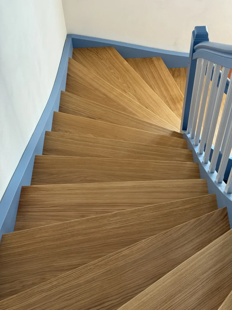 Blick die Wendeltreppe aus Holz hinunter, die hellblaue Zierleisten und ein weiß-blaues Geländer aufweist und vor einer hellen Wand steht.