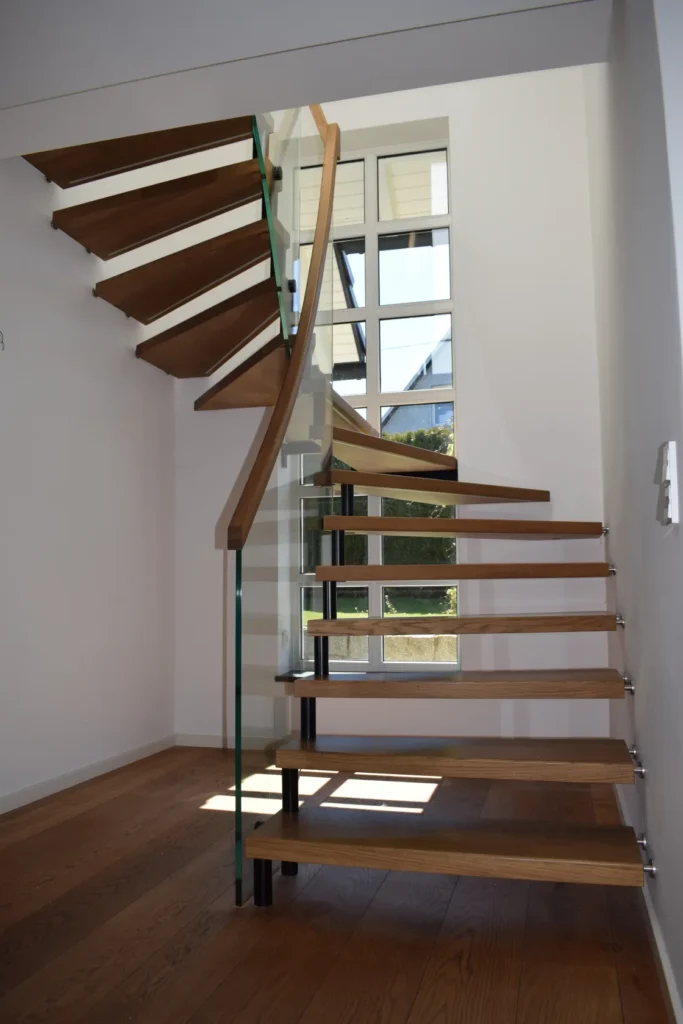 Eine moderne Holztreppe mit offenen Setzstufen und Glasgeländern schlängelt sich neben einem großen Fenster nach oben und lässt natürliches Licht in den Raum mit dem Holzboden strömen.