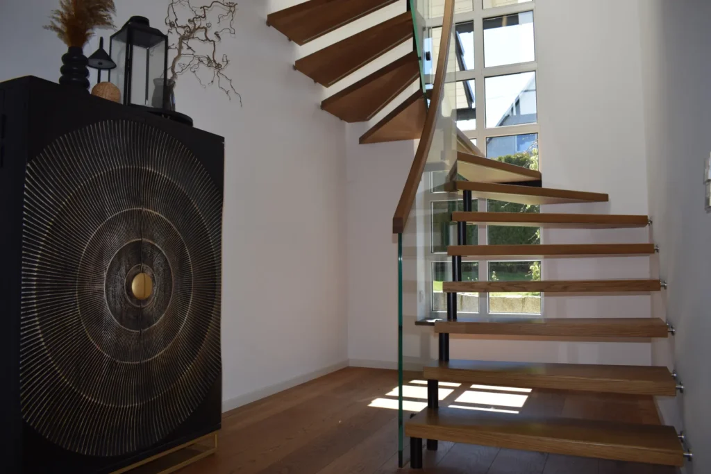 Modernes Interieur mit einer freitragenden Holztreppe und einem Glasgeländer, einem großen Fenster, das Tageslicht hereinlässt, sowie einem dekorativen schwarzen Schrank mit einem metallischen kreisförmigen Muster auf der linken Seite.