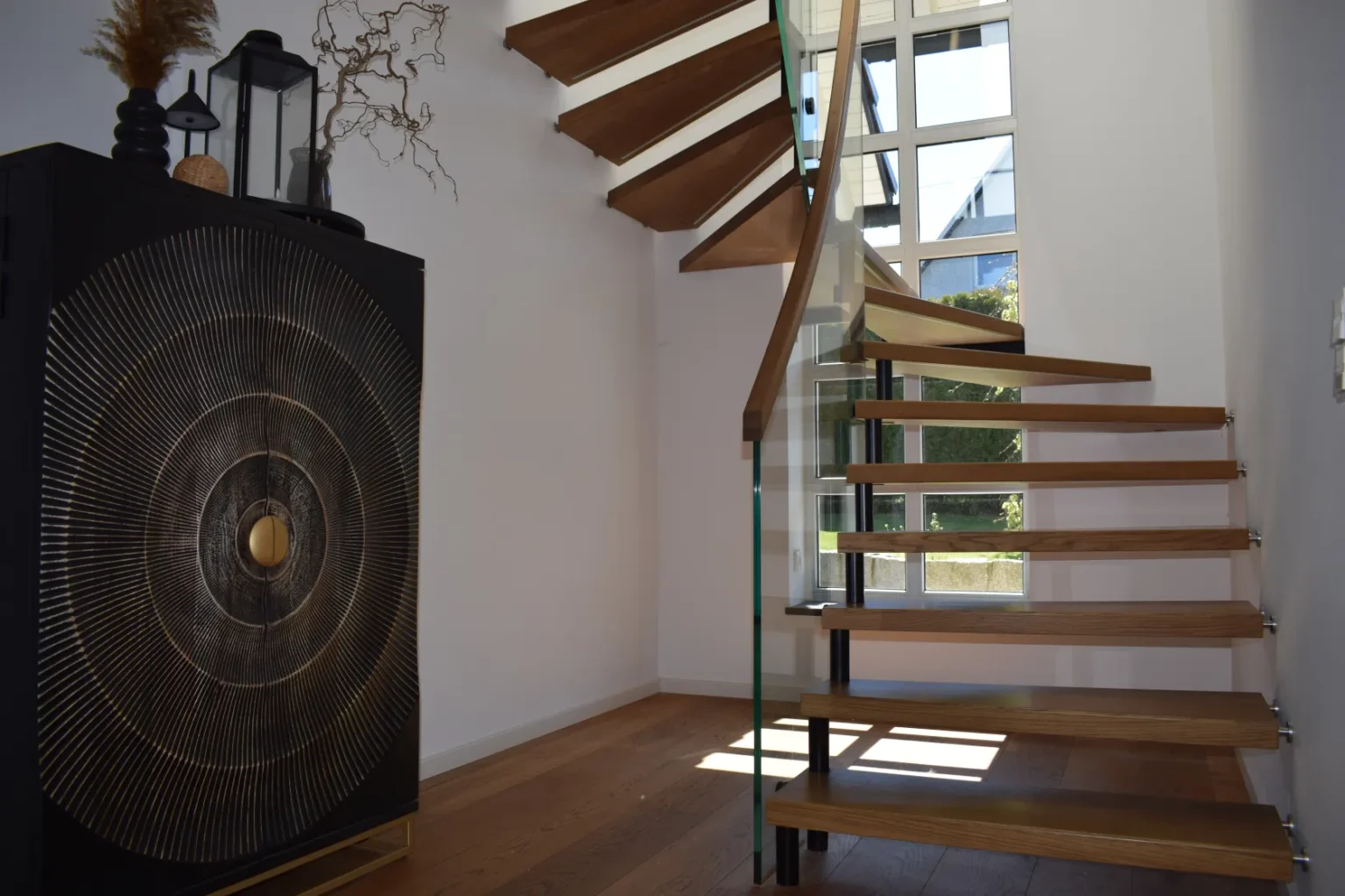 Modernes Interieur mit einer freitragenden Holztreppe und einem Glasgeländer, einem großen Fenster, das Tageslicht hereinlässt, sowie einem dekorativen schwarzen Schrank mit einem metallischen kreisförmigen Muster auf der linken Seite.