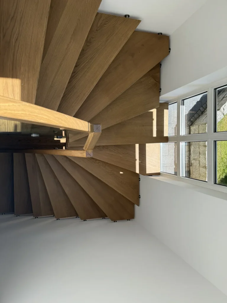 Eine spiralförmige Holztreppe mit breiten Stufen, von oben gesehen, neben einer weißen Wand und großen Fenstern, durch die Tageslicht hereinströmt.
