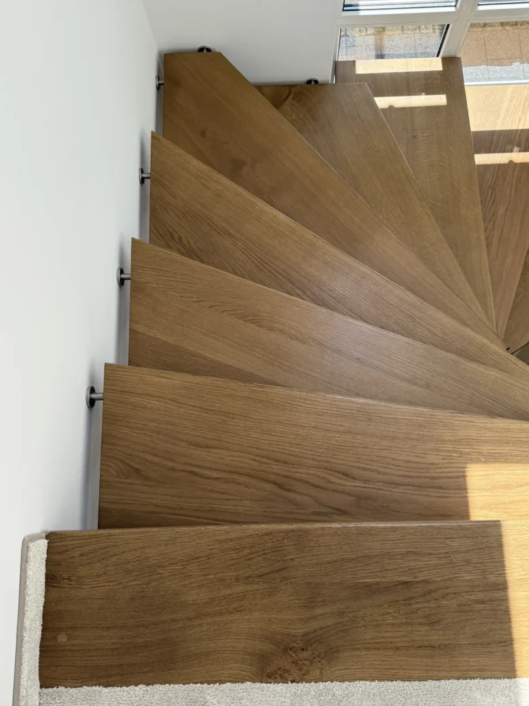 Eine Ansicht von oben auf eine moderne Holztreppe mit dreieckigen Stufen, die vom Sonnenlicht beleuchtet wird, das durch die nahegelegenen Fenster fällt. An der weißen Wand links sind Metallstützen befestigt, die die Treppe stützen.