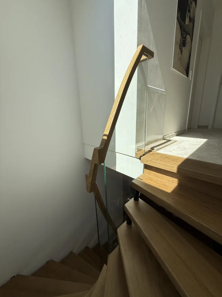 Eine moderne Holztreppe mit Glaseinsätzen schlängelt sich nach oben und wird durch das natürliche Licht eines Fensters beleuchtet. Der obere Flur ist mit einem hellen Teppichboden ausgelegt, und an der Wand hängt ein Schwarz-Weiß-Foto.