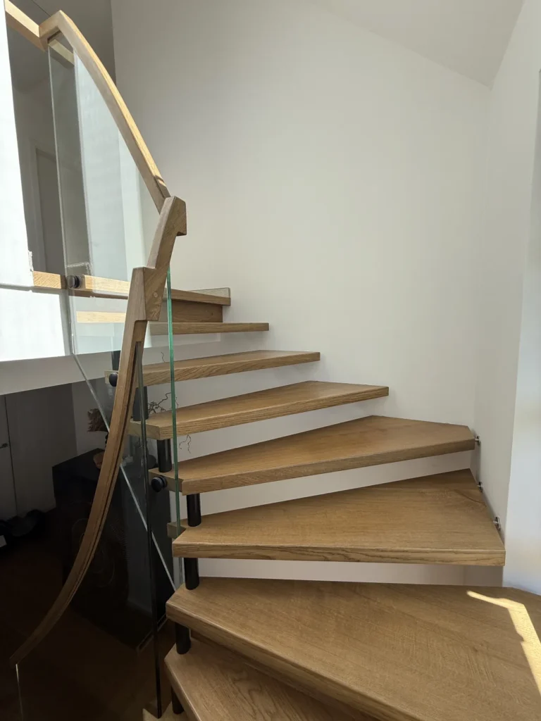 Eine moderne Holztreppe mit offenen Setzstufen und Glasgeländer schlängelt sich an einer weißen Wand empor und lässt so das natürliche Licht auf die Stufen fallen.