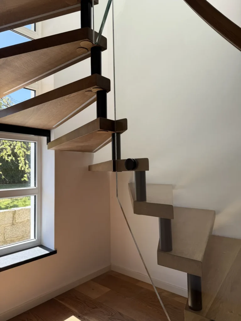Eine moderne Holztreppe mit offenen Setzstufen und Metallstützen, neben einem großen Fenster, durch das Sonnenlicht hereinströmt und Schatten auf die weißen Wände und den Holzboden wirft.