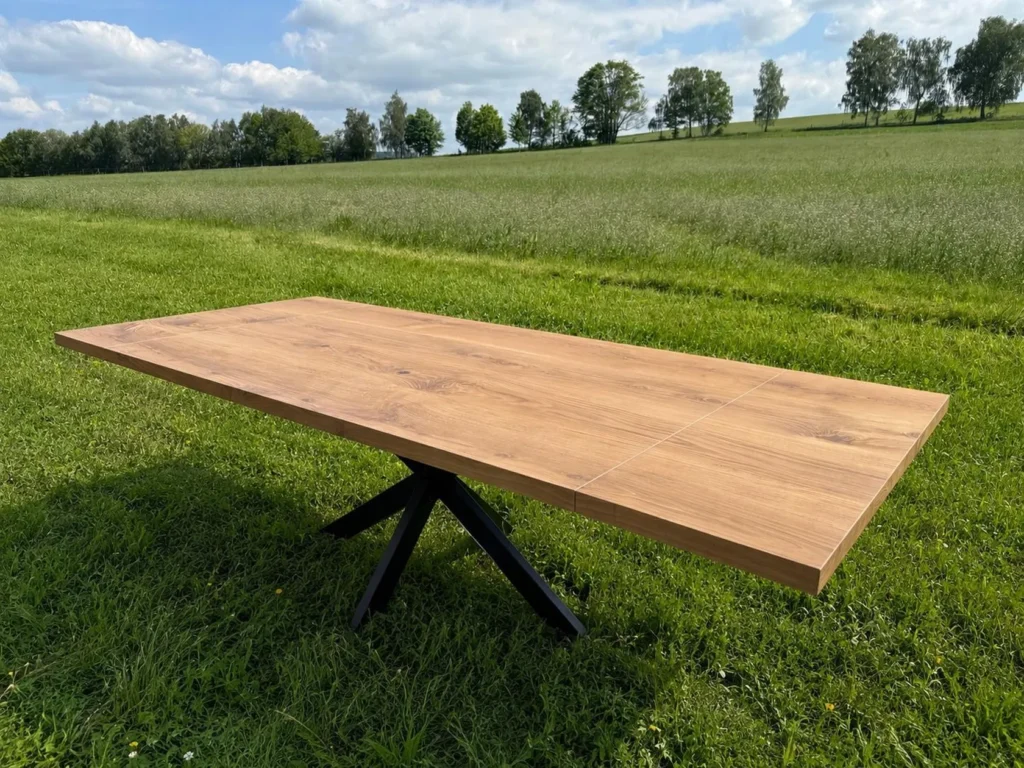 Ein großer Holztisch mit schwarzen Metallbeinen steht auf grünem Gras auf einer offenen Wiese, im Hintergrund sind Bäume und ein bewölkter blauer Himmel zu sehen.