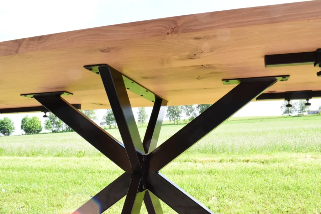 Nahaufnahme der Unterseite eines Holztisches mit schwarzen Metallbeinen, die ein X bilden; der Tisch steht im Freien auf einer Wiese, im Hintergrund sind Bäume zu sehen.