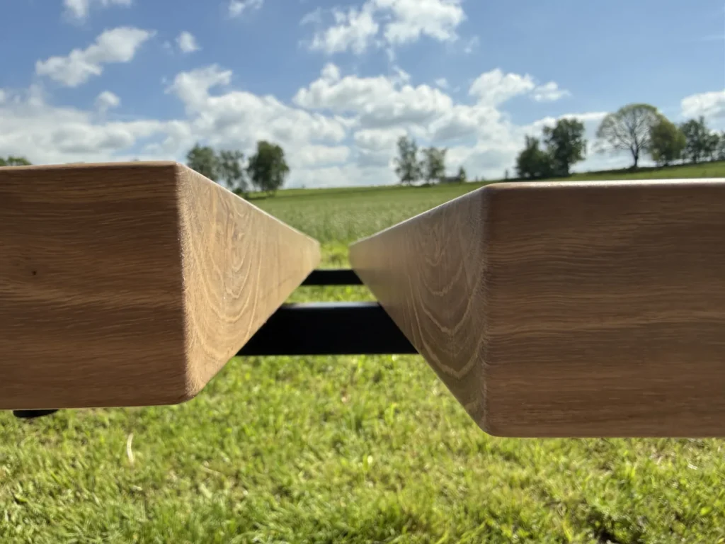 Nahaufnahme von zwei parallel verlaufenden Holzbalken, im Hintergrund eine Wiese, Bäume und ein teilweise bewölkter Himmel. Das Bild wurde aus einem niedrigen Blickwinkel aufgenommen, wobei der Fokus auf den Balken und der dahinter liegenden Landschaft liegt.