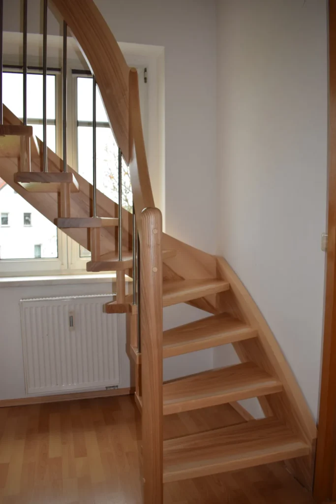 Eine moderne Holztreppe mit offenen Setzstufen und einem geschwungenen Handlauf, die neben einem Fenster hinaufführt. Die Treppe befindet sich über einem Heizkörper auf einem hellen Holzboden in einem hellen Raum.