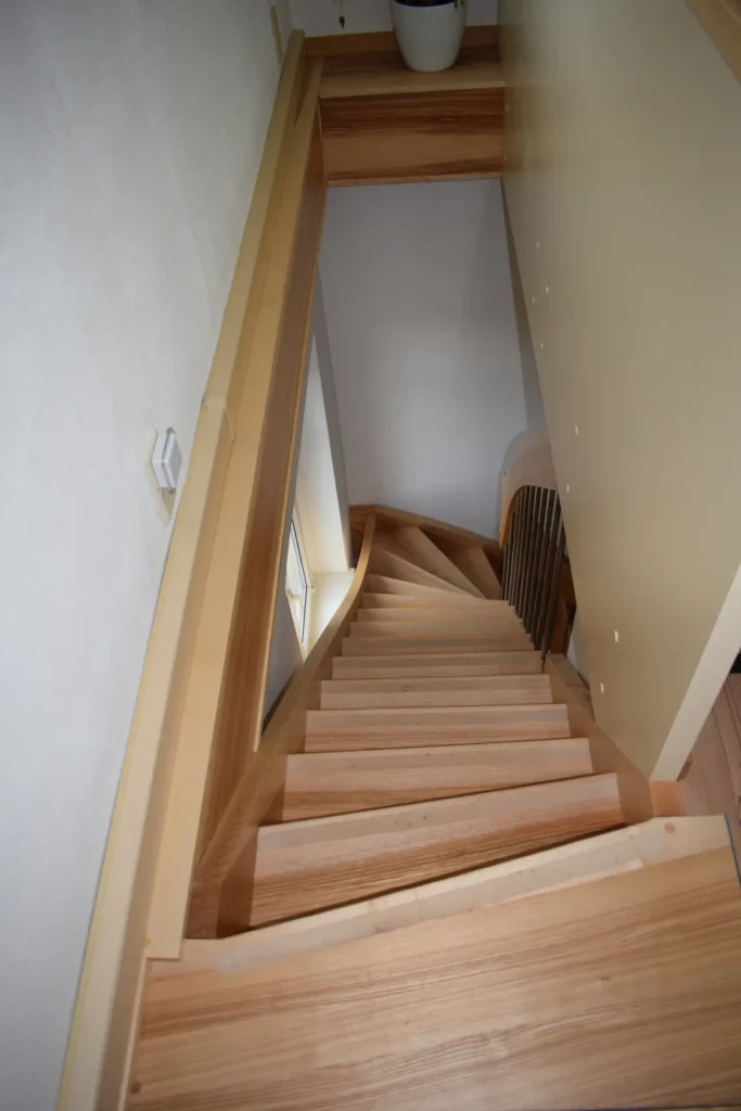 Eine schmale, steile Holztreppe mit schrägen Stufen und Geländern führt an einer hellen Wand entlang hinab und gelangt in einen kompakten Innenraum auf einer tiefer gelegenen Etage.