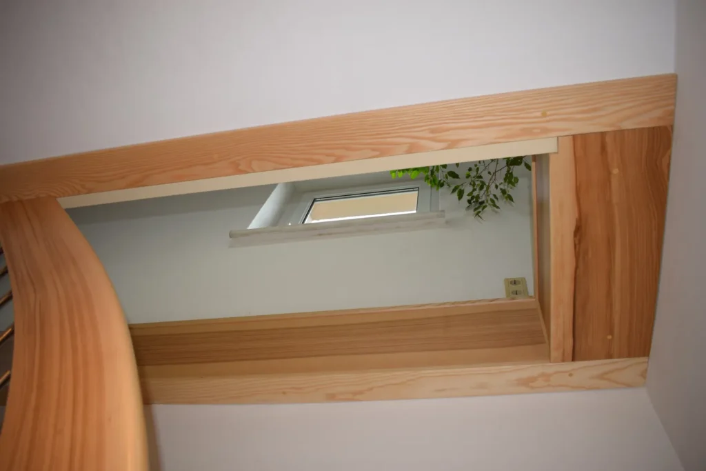 Blick von unten auf eine Holztreppe, der ein rechteckiges Fenster mit weißem Rahmen an einer hellen Wand zeigt; durch das Fenster sind einige grüne Blätter zu sehen, und sanftes Tageslicht fällt herein.