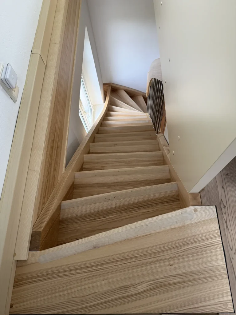 Ein Blick hinunter auf eine schmale Holztreppe mit ungewöhnlichen, unebenen Stufen und schräg verlaufenden Geländern, die sich durch natürliche Holztöne auszeichnet und am Fuß der Treppe ein Fenster aufweist.
