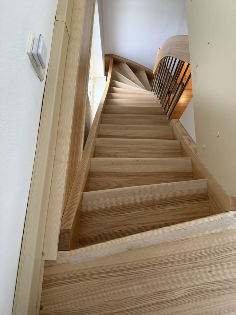 Eine Ansicht von oben auf eine schmale, spiralförmige Holztreppe mit hellen Holzstufen und einem Geländer auf der rechten Seite, neben einer weißen Wand und einem Fenster.