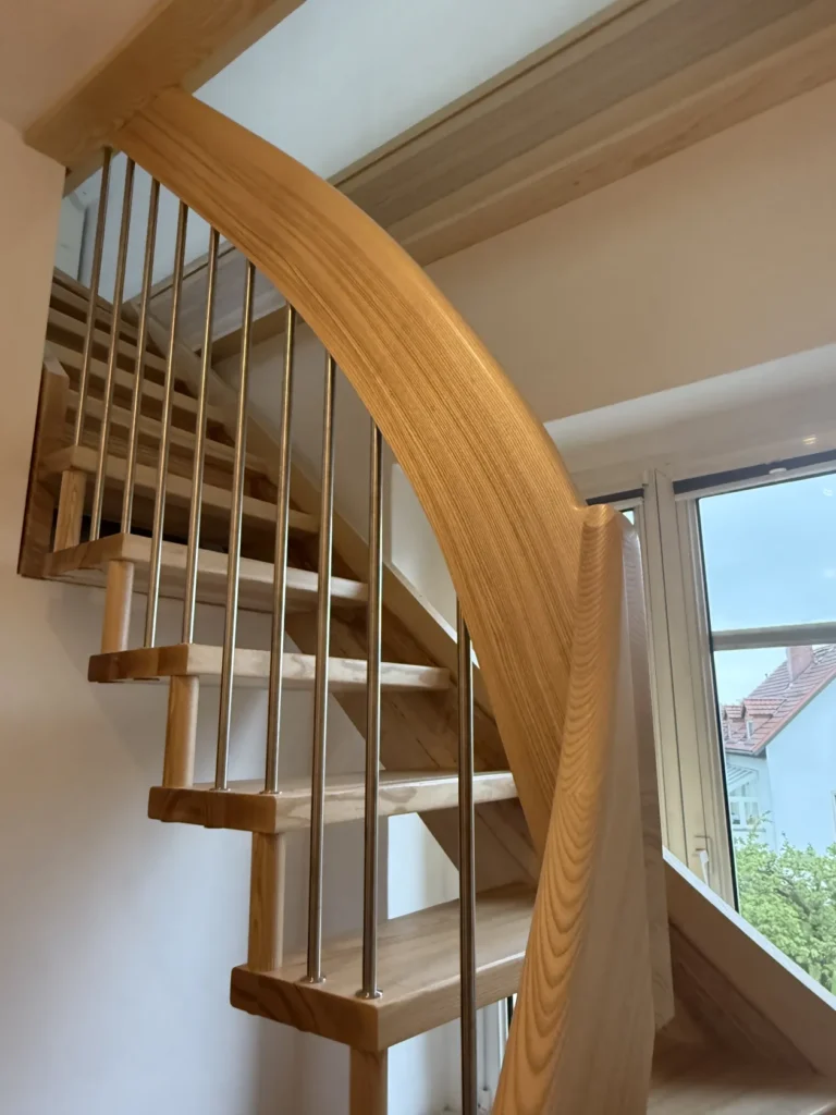 Eine moderne Holzwendeltreppe mit Metallgeländer, die sich durch sanfte Kurven und eine natürliche Holzoptik auszeichnet und zu einem hellen Bereich mit großen Fenstern und Blick auf die Dächer der umliegenden Häuser führt.