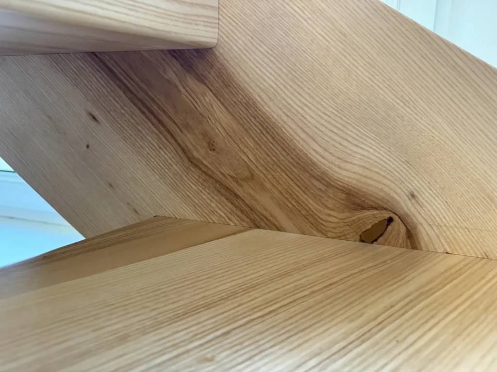 Nahaufnahme einer hellen Holztreppe, auf der die natürliche Holzmaserung und ein Ast zu sehen sind; drei Stufen sind erkennbar.
