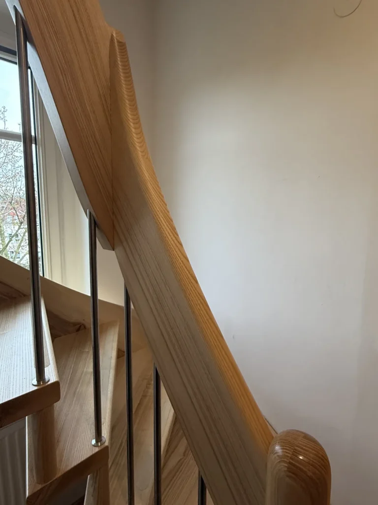 Eine Nahaufnahme einer modernen Holztreppe mit Metallgeländern, die den glatten, polierten Handlauf und die Stufen zeigt, neben einer weißen Wand und einem Fenster, durch das man Bäume sehen kann.
