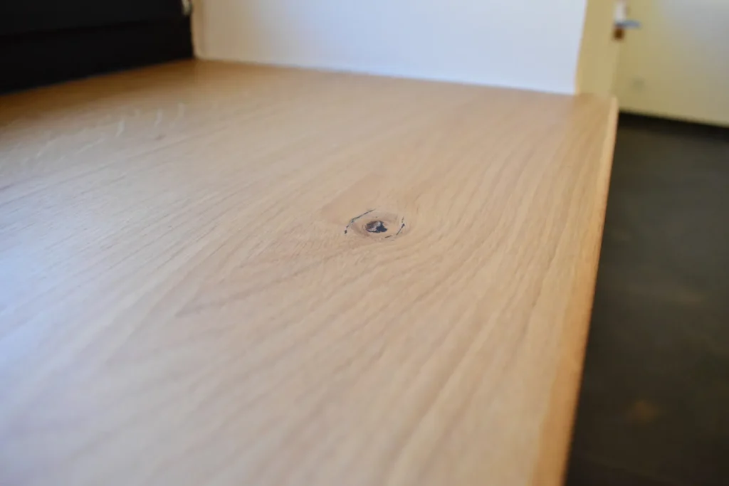 Nahaufnahme einer glatten, hellen Holzoberfläche mit einem sichtbaren Ast in der Maserung, daneben eine weiße Wand und im Hintergrund ein leicht unscharfer dunkler Boden sowie eine Tür.