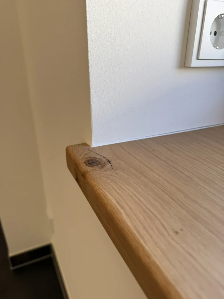 Eine Nahaufnahme einer Arbeitsplatte aus Holz mit einem sichtbaren Ast und einem Riss nahe dem Rand, neben einer weißen Wand und einer Steckdose.