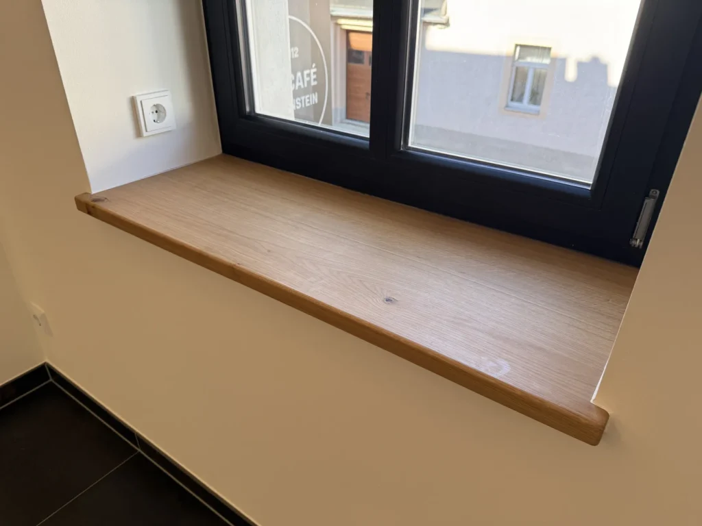 Unter einem schwarz gerahmten Fenster befindet sich eine Fensterbank aus Holz, an der linken Wand ist eine weiße Steckdose angebracht, und darunter liegen schwarze Bodenfliesen. Sonnenlicht fällt durch das Fenster herein und sorgt für eine sanfte Beleuchtung im Raum.
