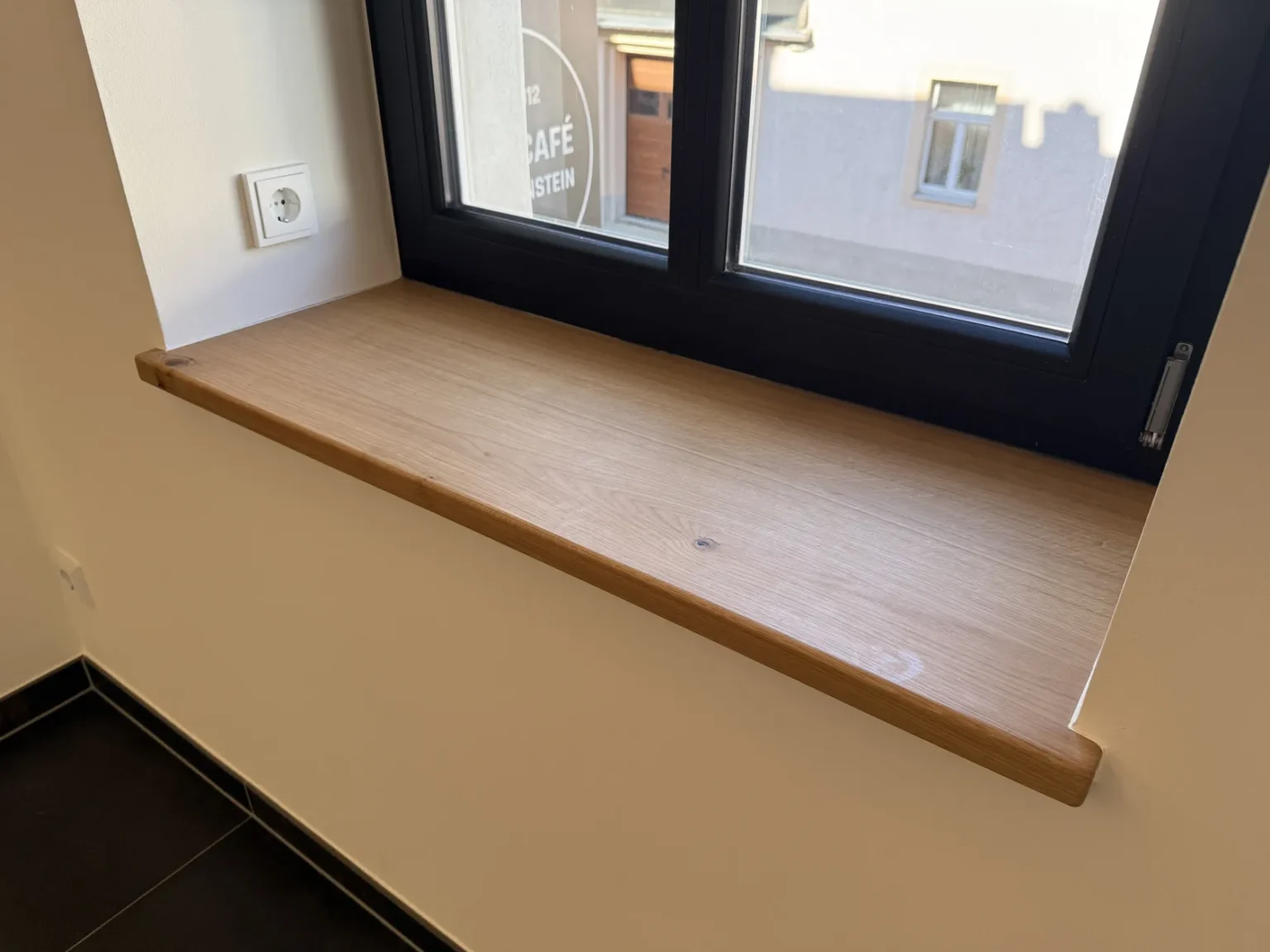 Unter einem schwarz gerahmten Fenster befindet sich eine Fensterbank aus Holz, an der linken Wand ist eine weiße Steckdose angebracht, und darunter liegen schwarze Bodenfliesen. Sonnenlicht fällt durch das Fenster herein und sorgt für eine sanfte Beleuchtung im Raum.