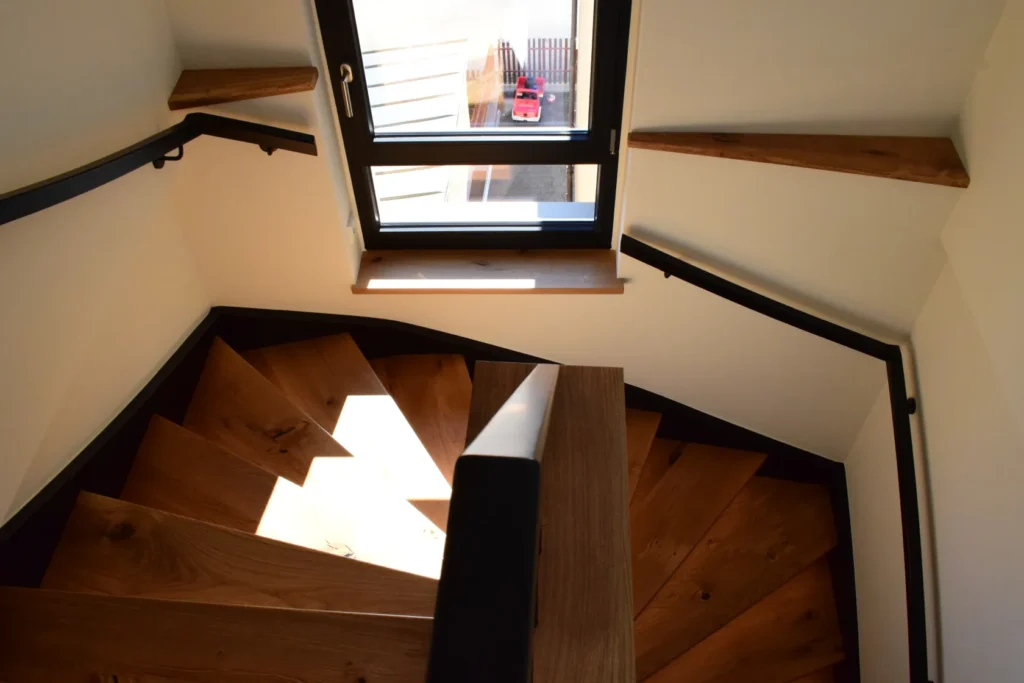 Eine Ansicht von oben auf eine moderne Holztreppe mit dunklen Handläufen, die vom Sonnenlicht durch ein großes Fenster beleuchtet wird und hinunter zu einem Fliesenboden führt, auf dem draußen ein kleines rotes Spielzeugauto zu sehen ist.