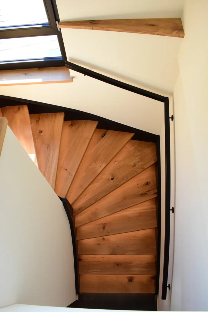 Eine Ansicht von oben auf eine moderne Holzwendeltreppe mit hellbraunen Stufen, schwarzem Geländer und weißen Wänden. Sonnenlicht fällt durch ein Fenster herein, wirft Schatten und beleuchtet einen Teil der Treppe.