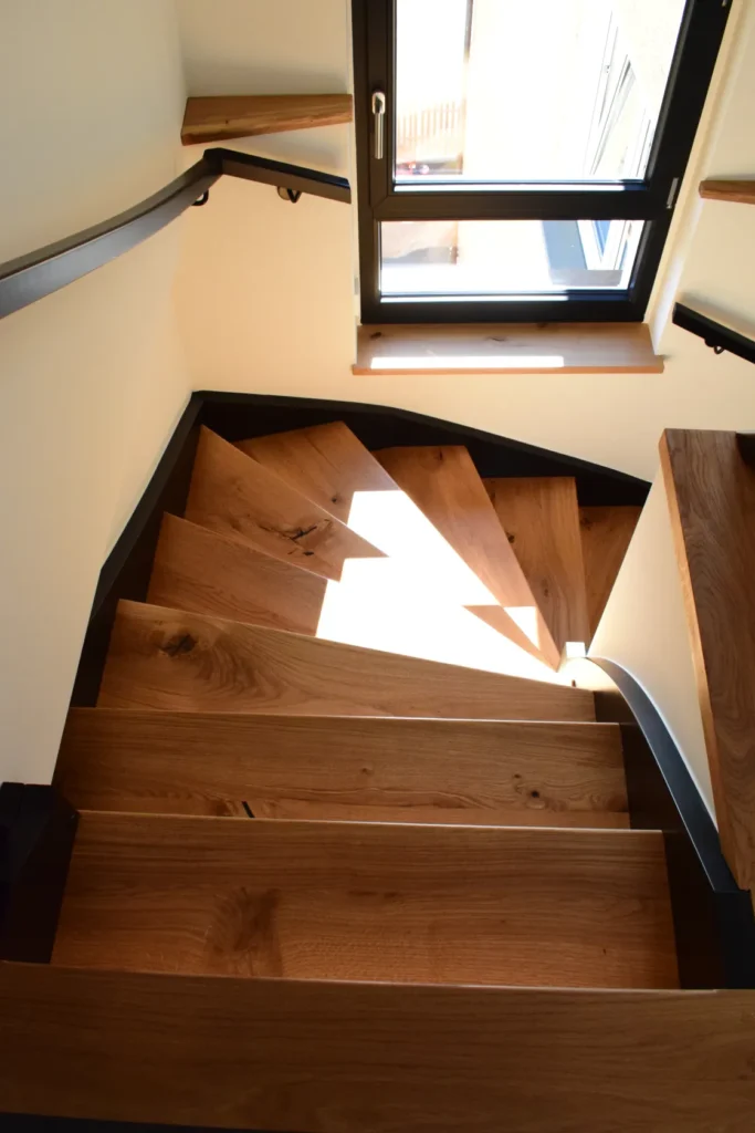 Eine spiralförmige Holztreppe mit dunklen Zierleisten, die durch natürliches Sonnenlicht aus einem Fenster im oberen Bereich beleuchtet wird. Die Treppe windet sich eng nach oben, und das warme Licht bringt die Holzmaserung besonders gut zur Geltung. Ein an der Wand befestigter Handlauf folgt der Kurve der Treppe.