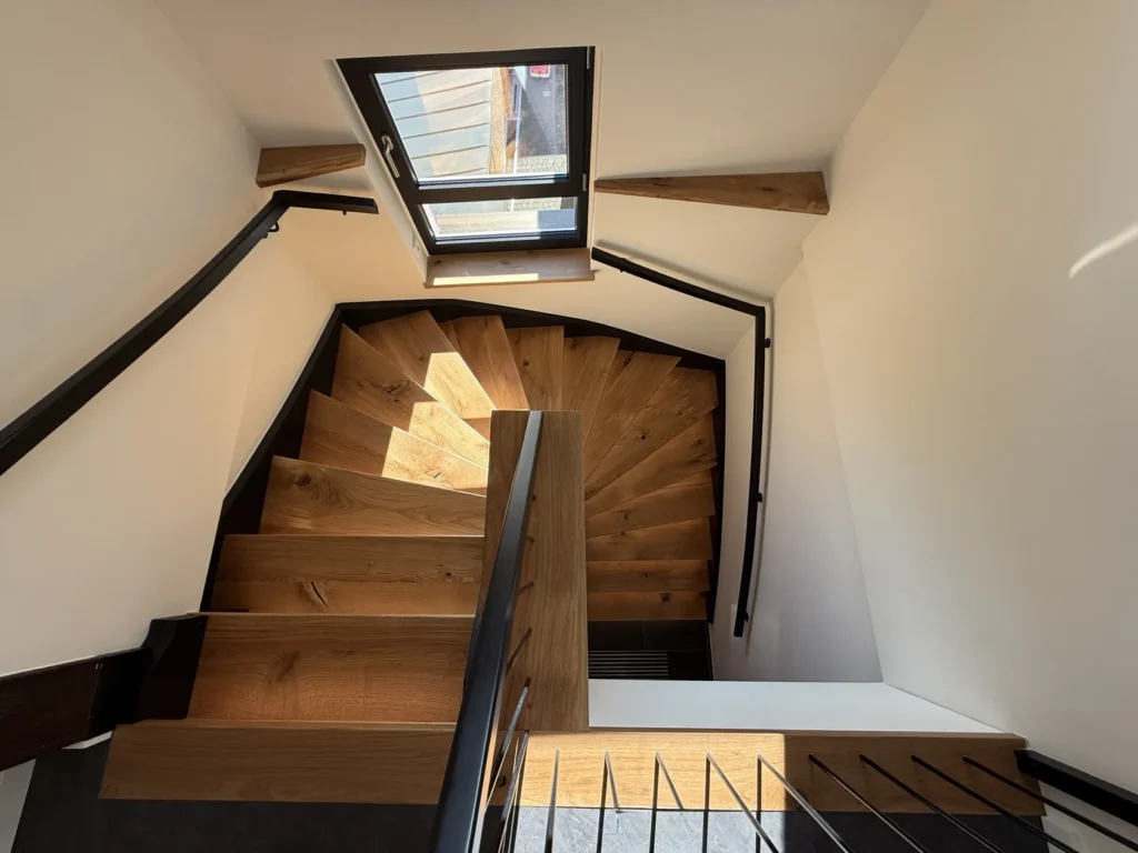 Eine Ansicht von oben auf eine moderne Holzwendeltreppe mit schwarzen Geländern, die von Sonnenlicht beleuchtet wird, das durch ein Fenster am oberen Ende der Treppe hereinströmt. Die Stufen bilden ein dynamisches, geometrisches Muster.