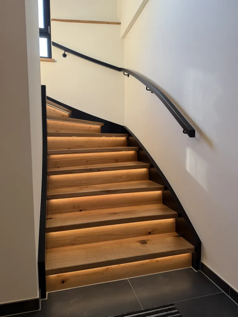 Eine moderne Holztreppe mit integrierter, warmer LED-Beleuchtung unter jeder Stufe, schwarzen Handläufen und weißen Wänden, die sich nach oben zu einem Fenster schlängelt, durch das Tageslicht hereinströmt.