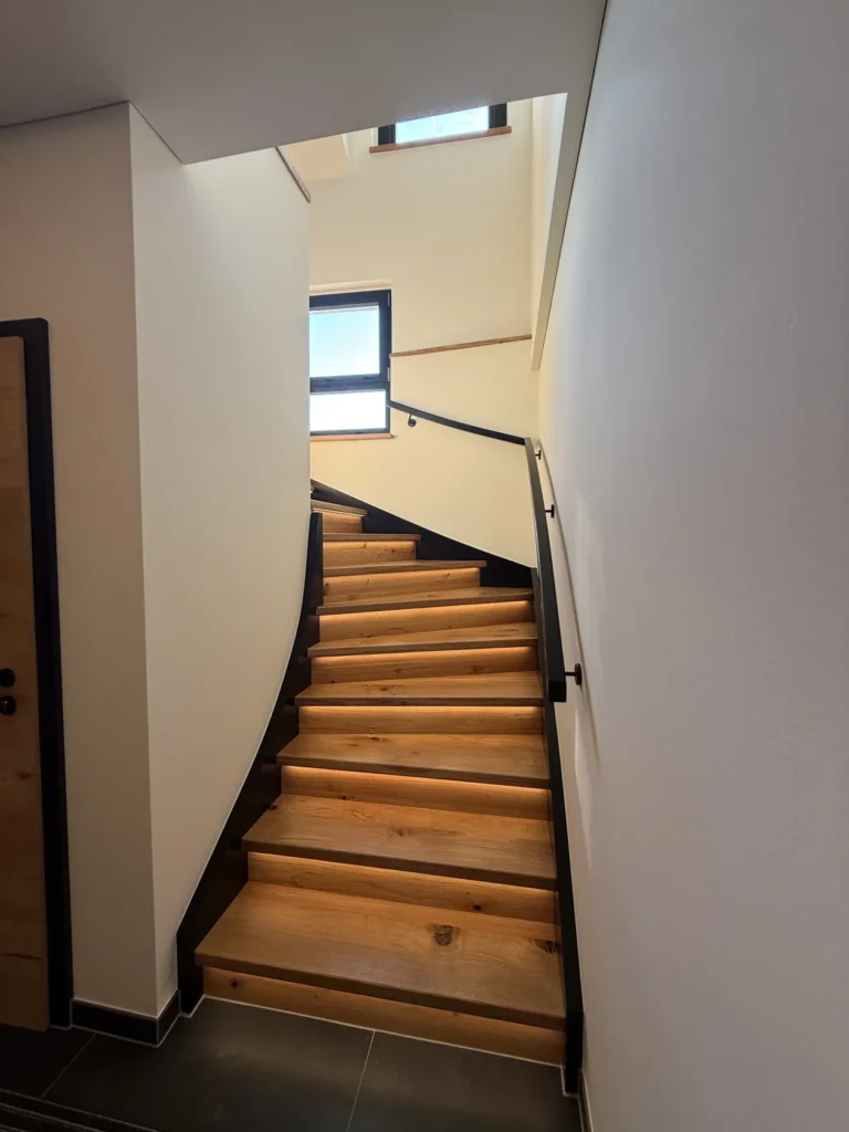 Eine moderne Innentreppe mit Holzstufen und schwarzen Metallgeländern schlängelt sich zwischen weißen Wänden nach oben und führt zu einem hell beleuchteten Obergeschoss mit zwei Fenstern.