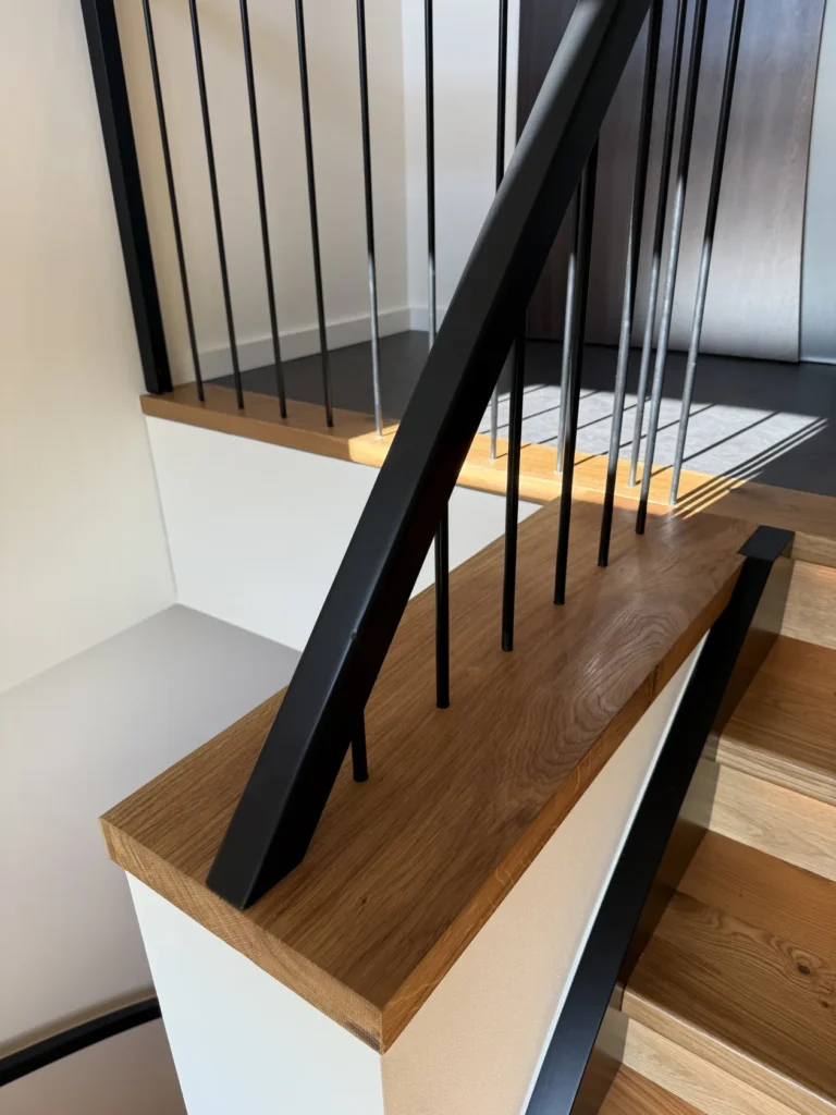 Eine Nahaufnahme einer modernen Holztreppe mit schwarzen vertikalen Metallgeländern und einem Handlauf; Sonnenlicht fällt durch ein nahegelegenes Fenster auf den dunklen Boden am oberen Ende der Treppe.