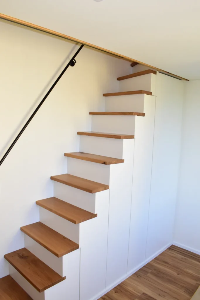 Eine moderne, schmale Holztreppe mit weißen Seitenwänden und offenen Setzstufen führt an einer weißen Wand entlang nach oben. Sie verfügt über einen schlichten schwarzen Handlauf und Stufen aus hellem Holz, unter denen Stauschränke eingebaut sind.
