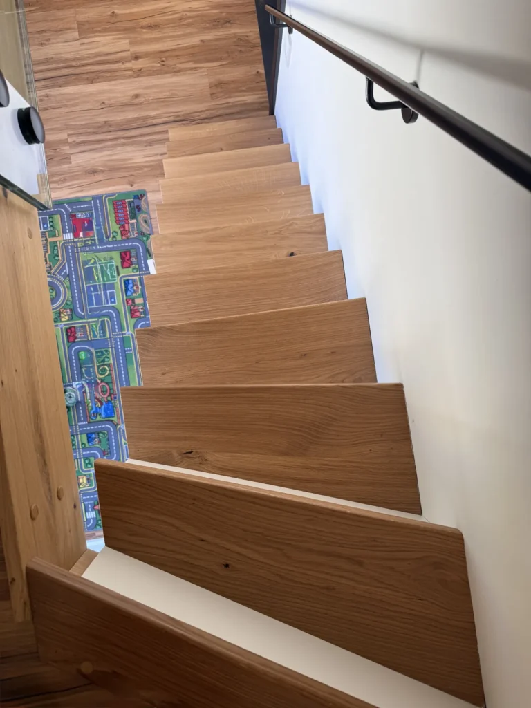 Eine Holztreppe mit abwechselnden Stufen führt hinunter zu einem Boden, der mit einer bunten Spielmatte bedeckt ist, auf der Straßen und Gebäude abgebildet sind. Ein schwarzer Handlauf verläuft entlang der weißen Wand auf der rechten Seite.