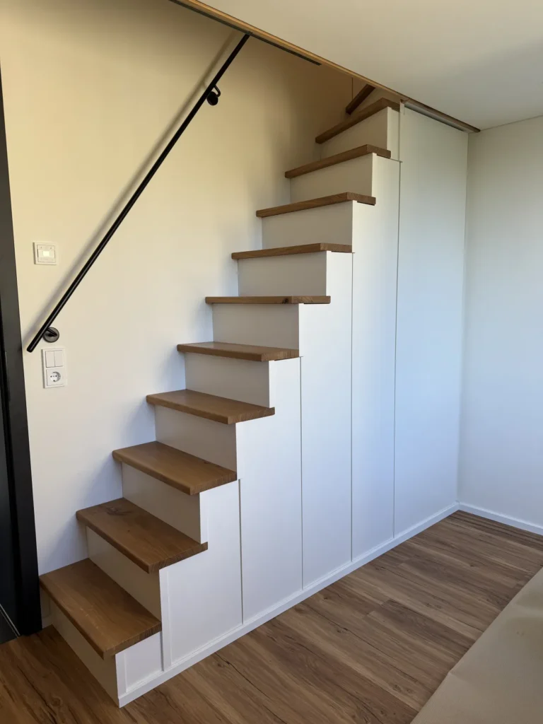 Eine moderne Holztreppe mit weißen Seitenwänden und einem schwarzen Handlauf führt in das Obergeschoss eines Raums mit Holzboden und weißen Wänden. Unter der Treppe sind Stauschränke eingebaut.