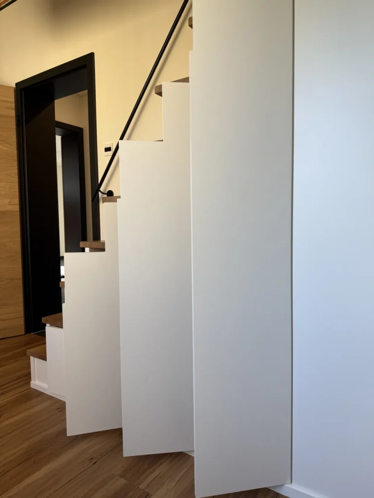 Eine moderne Treppe mit weißen, geometrischen Seitenwänden und Holzstufen, die vor einer Wand in Weiß mit Holzakzenten steht. Die Seitenwände bilden ein Zickzackmuster, und ein schwarzer Handlauf verläuft entlang der Treppe.