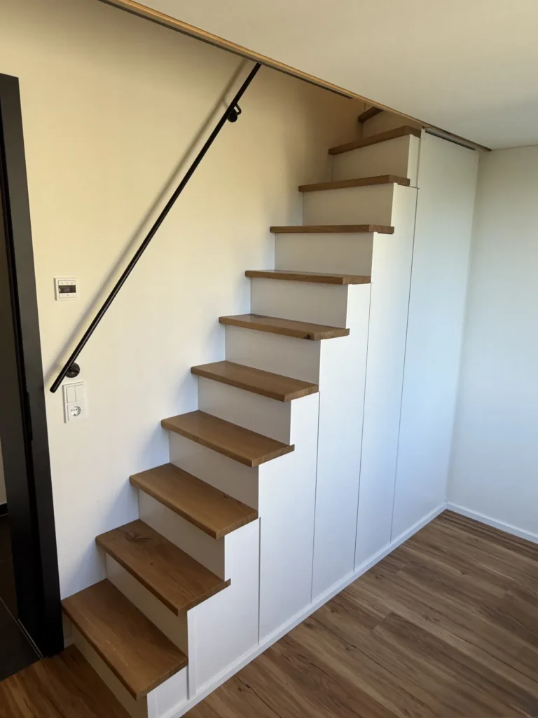 Eine moderne, steile Holztreppe mit weißen Seitenwänden und einem schwarzen Handlauf, die an einer weißen Wand angebaut ist; unter der Treppe befinden sich Aufbewahrungsschränke, und sie steht auf einem Holzboden.