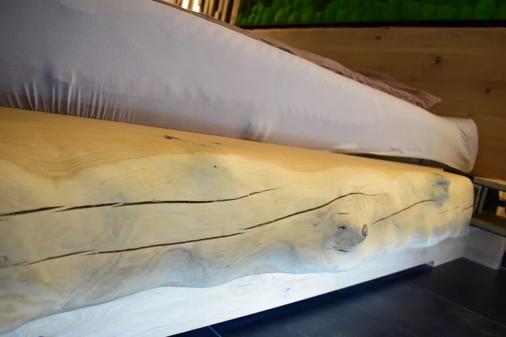 Eine Nahaufnahme eines Bettgestells aus Holz, das aus einem dicken, naturbelassenen Baumstamm mit sichtbaren Astlöchern und Maserung gefertigt ist und auf dem eine Matratze mit einem hellen Spannbetttuch liegt.