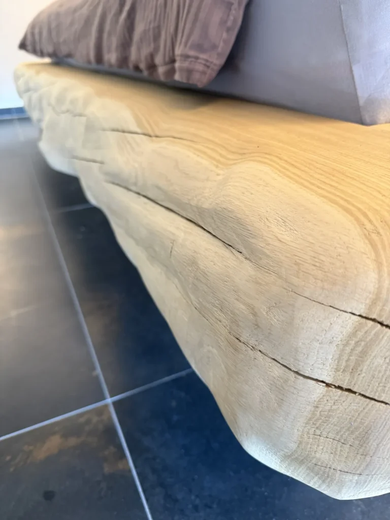 Nahaufnahme eines Bettes mit einem natürlichen, unbehandelten Holzrahmen, dessen Maserung und Risse zu sehen sind, das auf einem glänzenden, dunklen Fliesenboden steht. Am oberen Bildrand sind Kissen und Bettwäsche teilweise zu sehen.