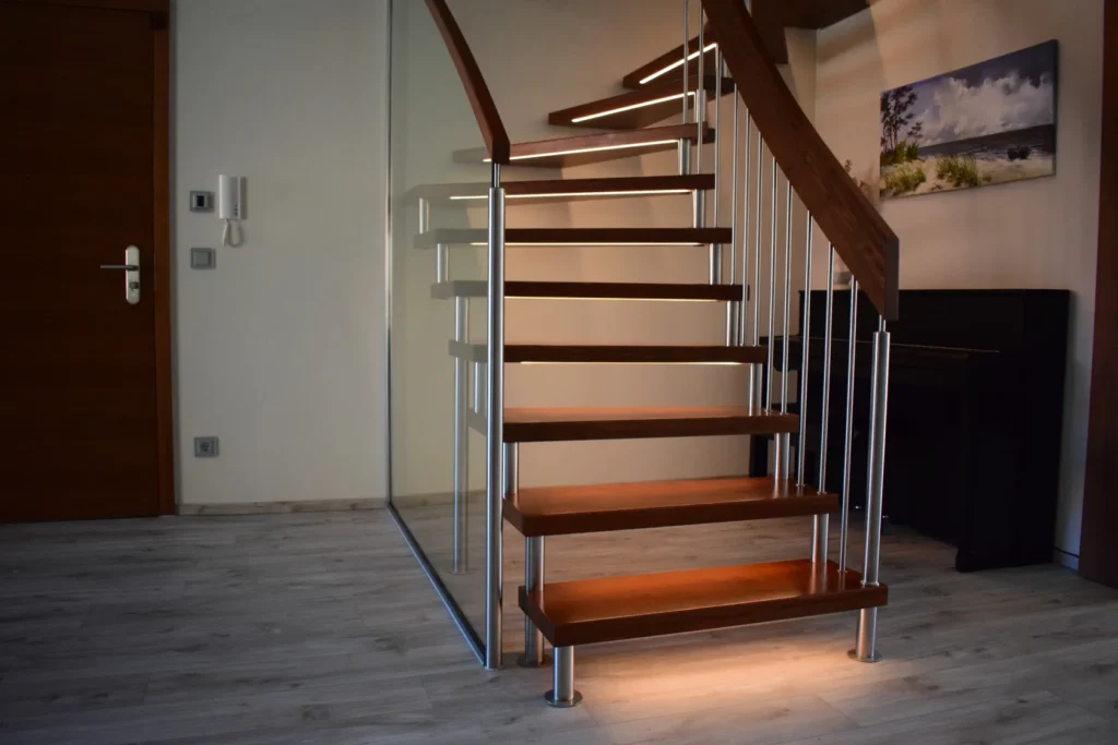 In einem hellen Raum mit hellem Holzboden, einer Glaswand, einem Klavier, einem Gemälde und einer geschlossenen Holztür steht eine moderne Holztreppe mit Metallgeländern und integrierter Beleuchtung.