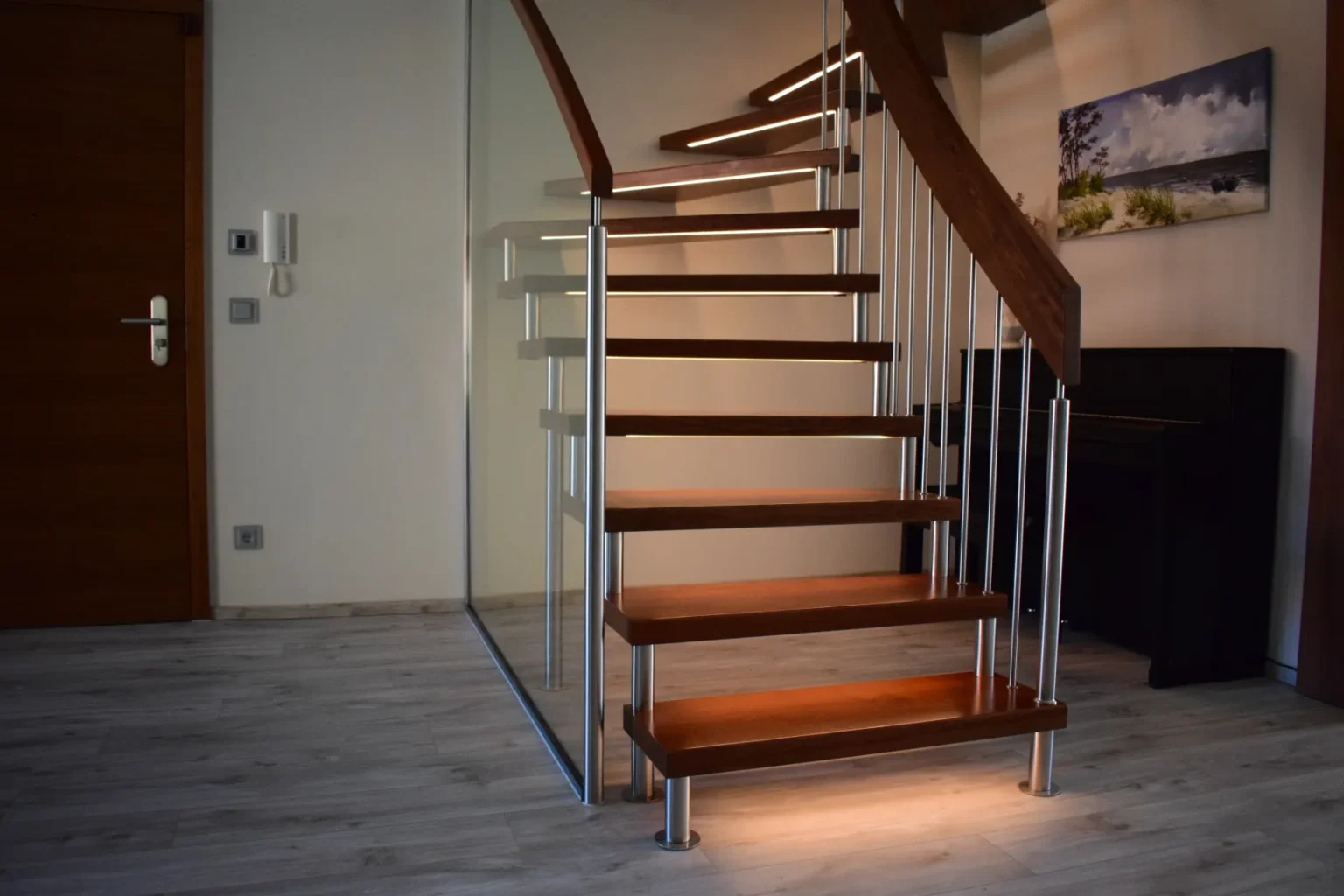 In einem hellen Raum mit hellem Holzboden, einer Glaswand, einem Klavier, einem Gemälde und einer geschlossenen Holztür steht eine moderne Holztreppe mit Metallgeländern und integrierter Beleuchtung.