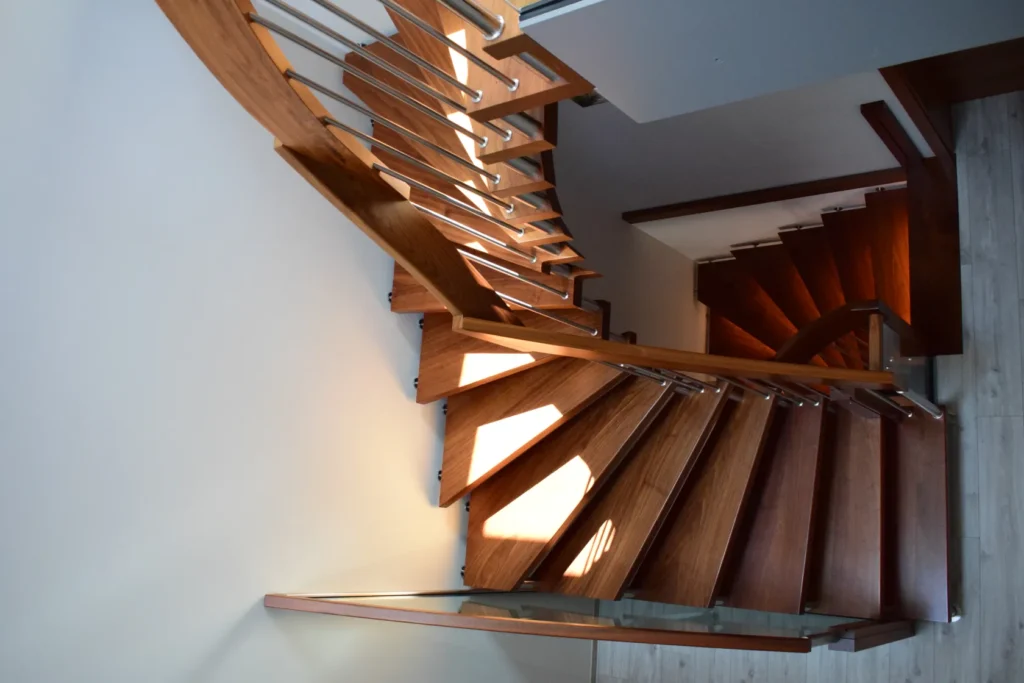 Eine Ansicht von oben auf eine moderne Holztreppe mit Metallgeländern, die sich geschwungen an einer hellen Wand entlang nach unten windet und auf einem hellen Holzboden endet. Das Sonnenlicht wirft Muster auf die Stufen.