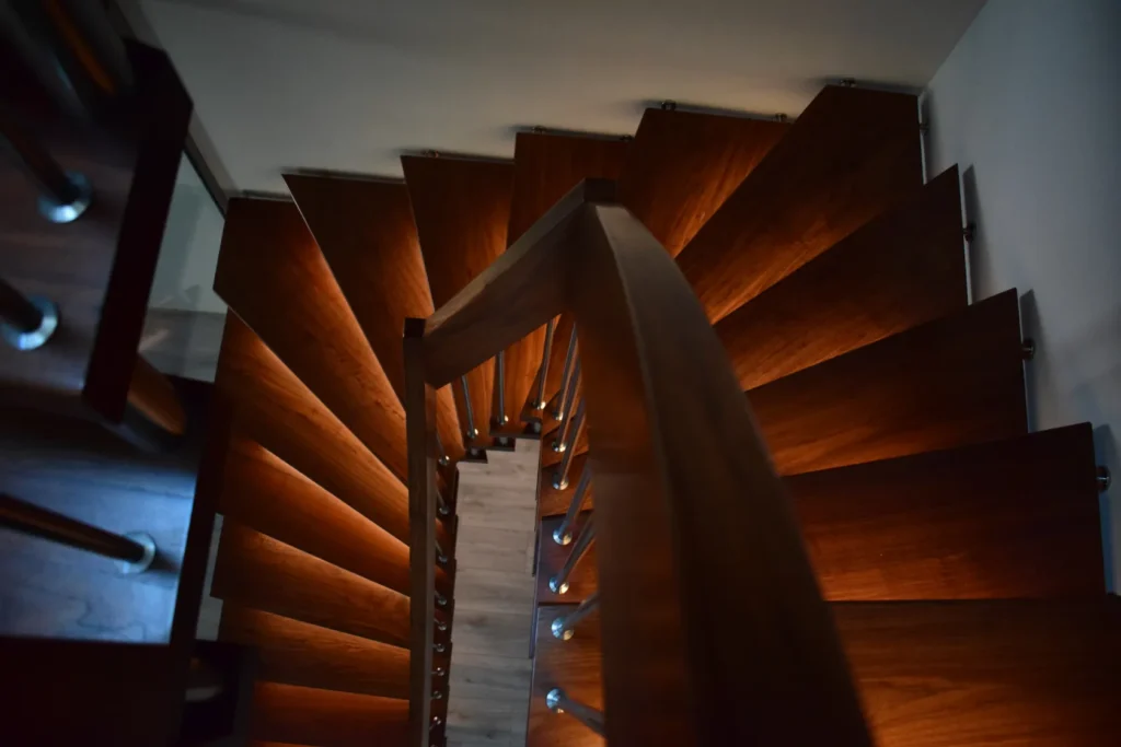 Eine Ansicht von oben auf eine spiralförmige Holztreppe, deren einzelne Stufen von warmer Beleuchtung erhellt werden, wodurch die satten Holztöne und das elegante, moderne Design besonders zur Geltung kommen.
