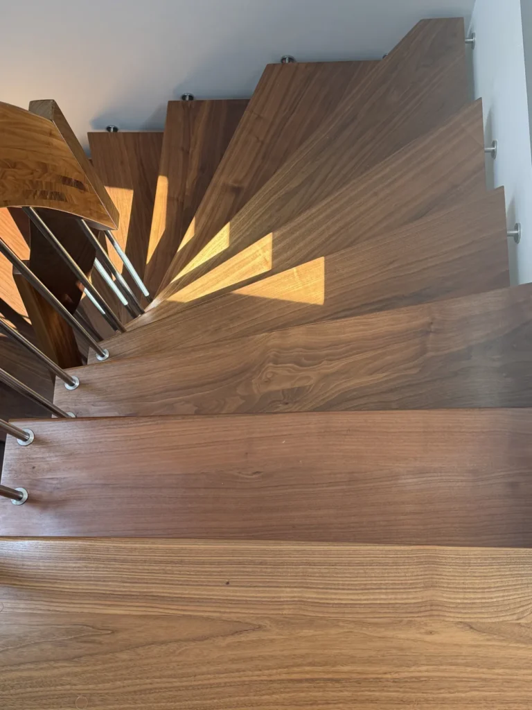 Eine Ansicht von oben auf eine moderne Holzwendeltreppe, auf deren Stufen das Sonnenlicht Schatten wirft, mit einem eleganten Geländer aus Metall und Holz auf der linken Seite.