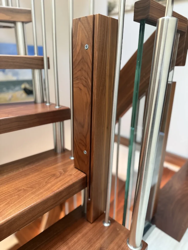 Nahaufnahme einer modernen Holztreppe mit Metallgeländer und einer Glasplatte, die die Holzmaserung und die Metallbefestigungen hervorhebt. Im Hintergrund ist ein unscharfes Gemälde zu sehen.