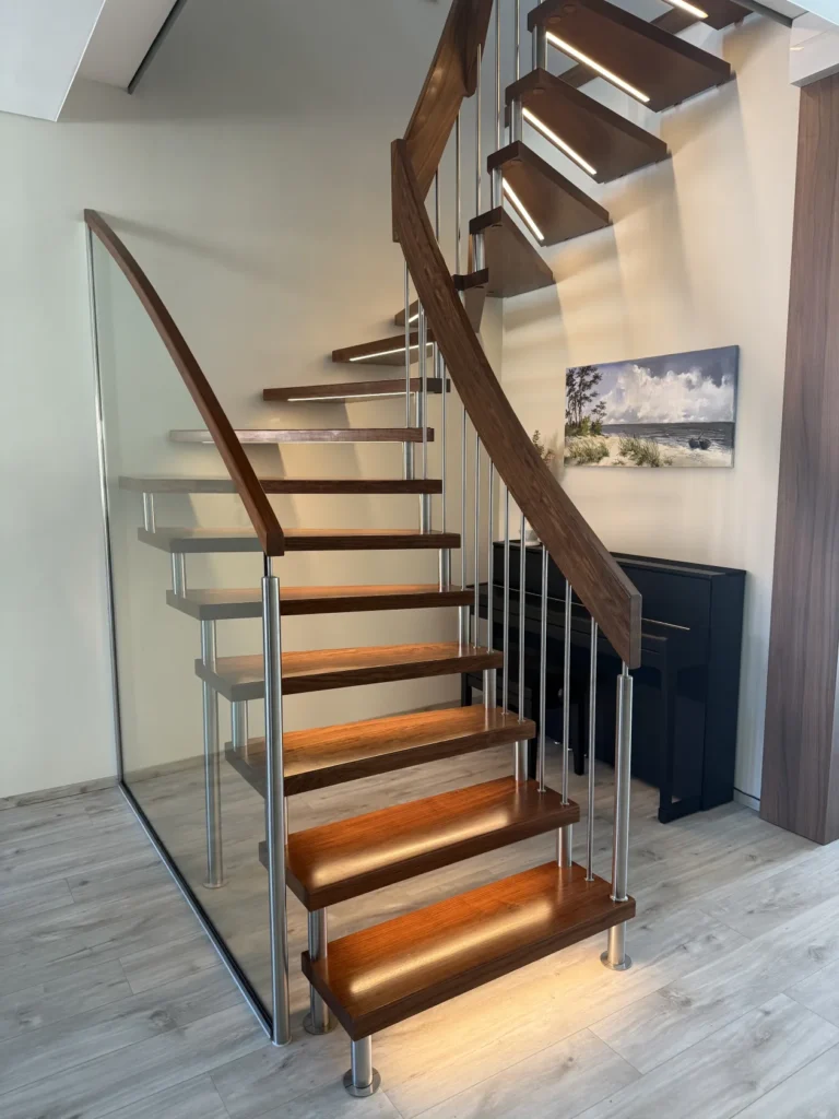 Eine moderne Holztreppe mit Metallgeländer schlängelt sich in einem hellen Raum mit hellen Holzböden nach oben. Unter der Treppe sind ein Klavier und ein Landschaftsgemälde zu sehen. Sanfte Beleuchtung setzt die Stufen in Szene.