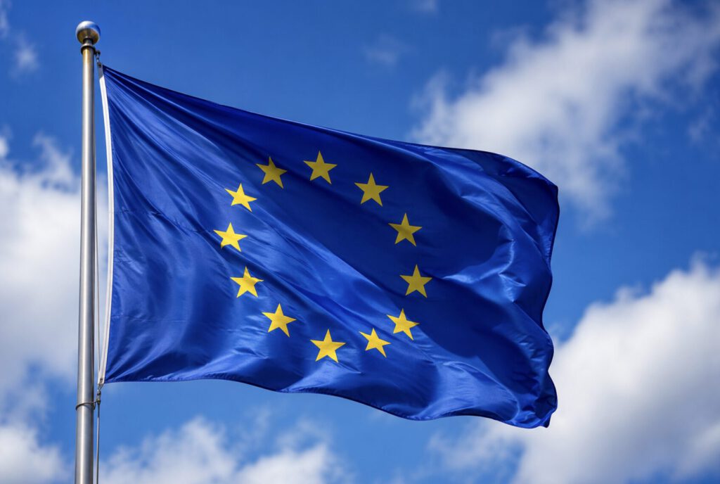 Die Flagge der Europäischen Union, auf der zwölf gelbe Sterne in einem Kreis auf blauem Hintergrund zu sehen sind, weht an einem Fahnenmast vor einem teilweise bewölkten blauen Himmel.