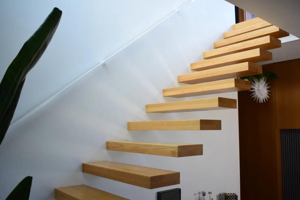 Eine an einer weißen Wand befestigte, freitragende Holztreppe führt in einem modernen, minimalistischen Interieur nach oben; daneben sind eine grüne Pflanze und zeitgenössische Dekorationselemente zu sehen.